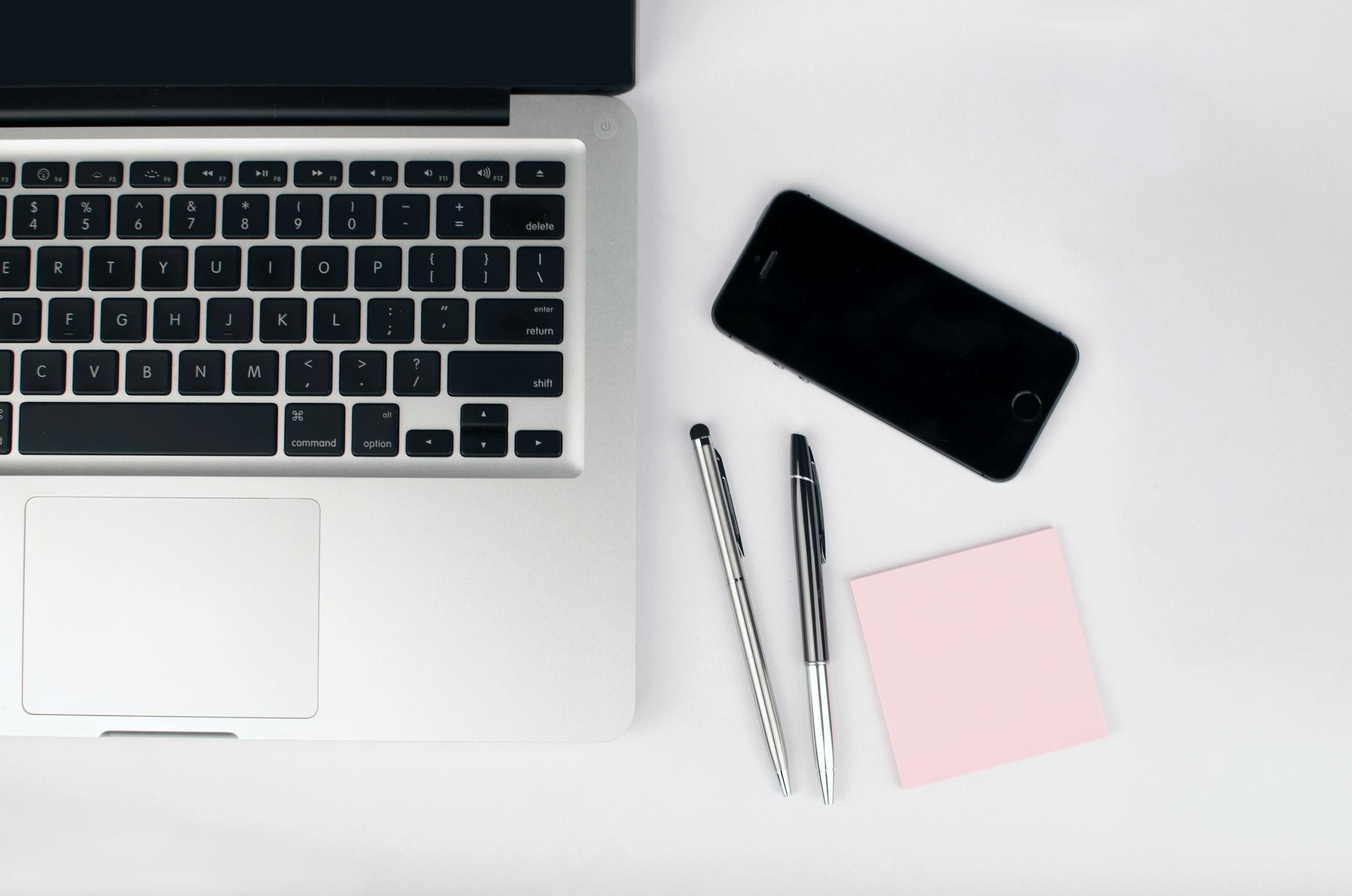 A top view of a modern workspace featuring a laptop, smartphone, pens, and a pink sticky note on a white desk.
